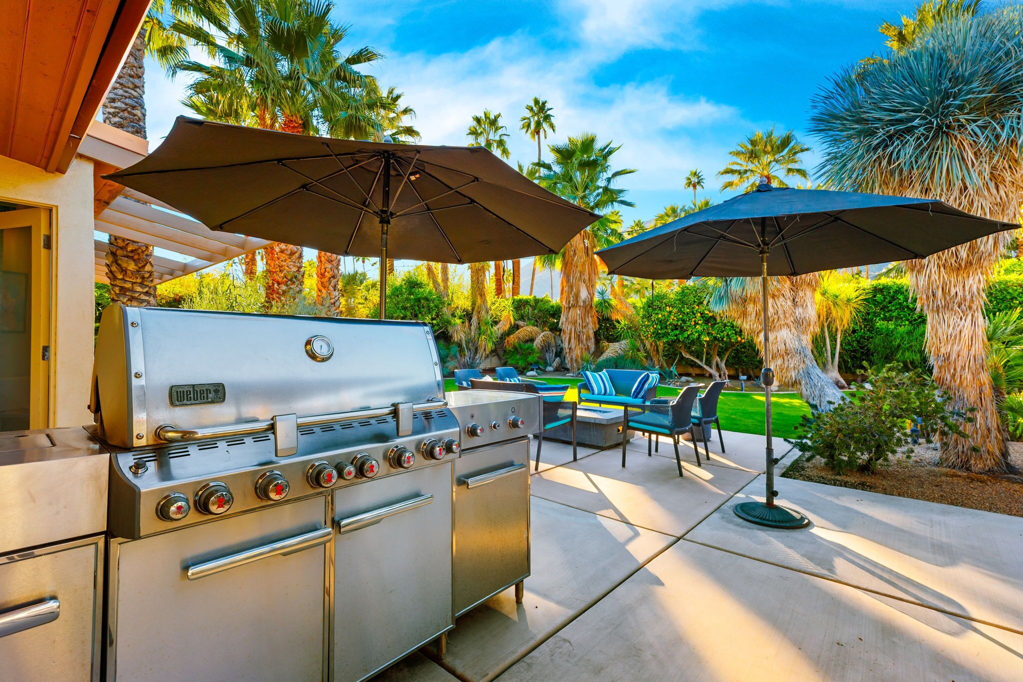 Een Weber barbecue met vier branders en een bijgelegen oven, op een terras met tuinmeubels en parasols.