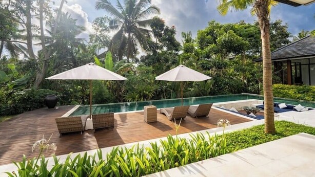 A poolside area with lounge chairs and umbrellas, surrounded by lush greenery and palm trees. - Villa Bae by GORO (Bali)