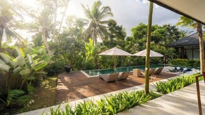 A pool area with lounge chairs and umbrellas, surrounded by lush greenery and palm trees. - Villa Bae by GORO (Bali)