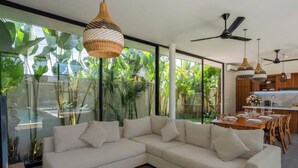 A modern living room with a white sectional sofa, large windows with tropical plants, and a dining area with wooden furniture. - Villa Bae by GORO (Bali)
