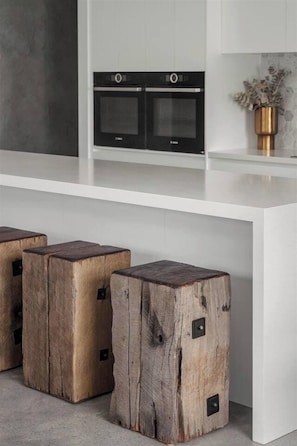 A modern kitchen with white cabinetry, two built-in ovens, and three wooden stools with metal accents.