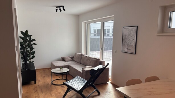 Ein modernes Wohnzimmer mit einer Eckcouch, einem runden Couchtisch, einem schwarzen Sessel und einem Esstisch. Ein großes Fenster sorgt für natürliches Licht und bietet einen Ausblick nach draußen.