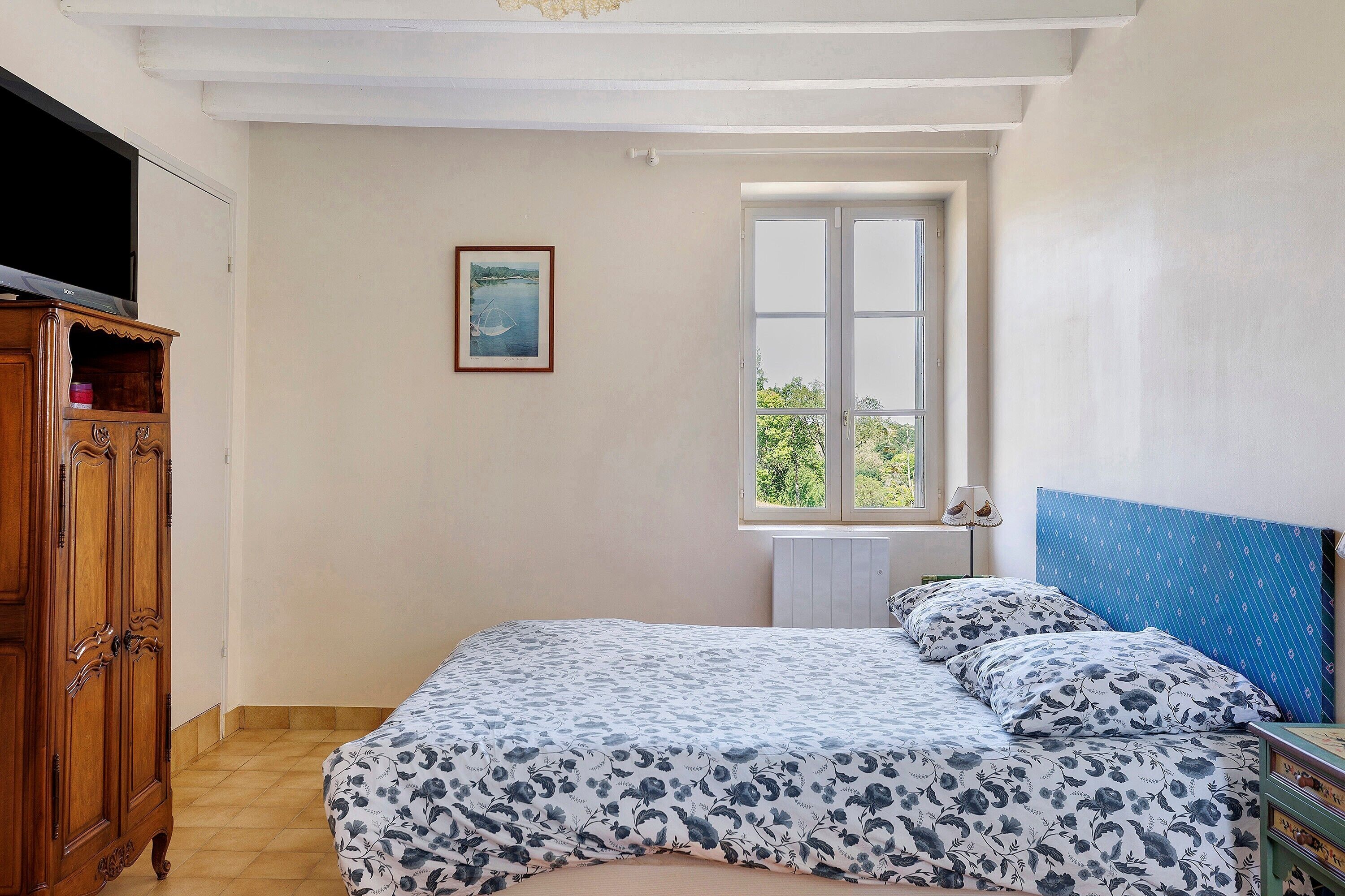 A bedroom with a bed, a window, a wooden wardrobe, and a framed picture on the wall.