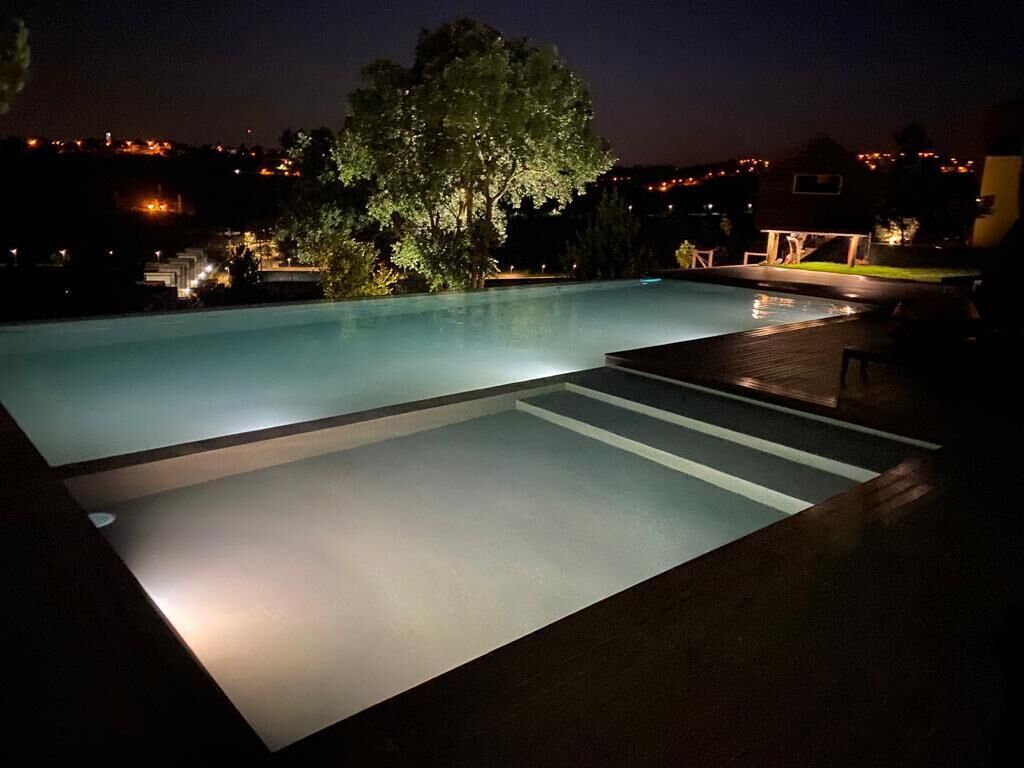 Une piscine illuminée la nuit, avec un arbre et une terrasse en bois.