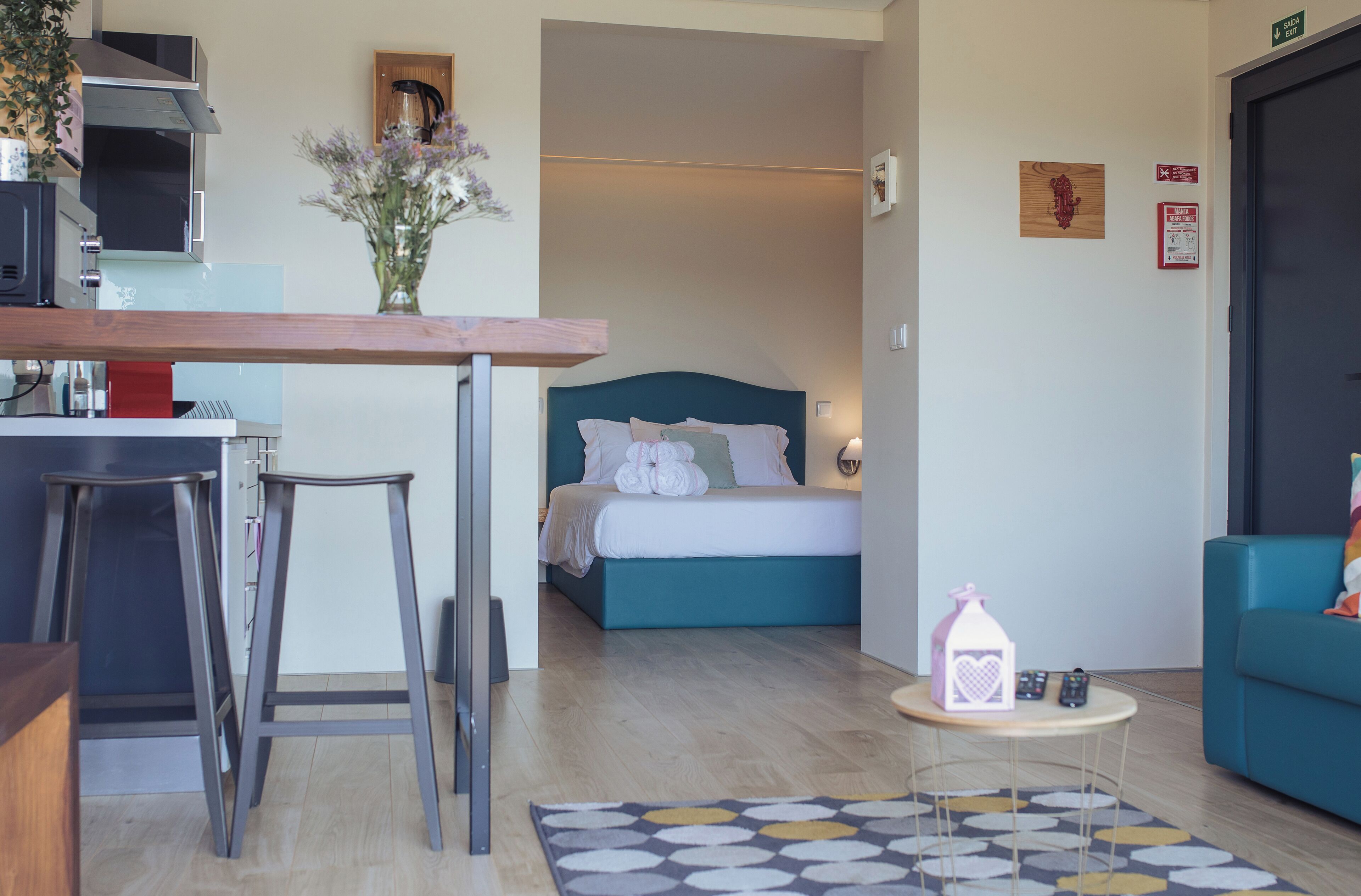 A modern hotel room with a kitchenette, a bed with a teal headboard, a small round table with a decorative item, and a blue sofa.