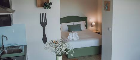 A compact hotel room with a kitchenette, a bed with green bedding, a bedside lamp, and a wall-mounted plant.