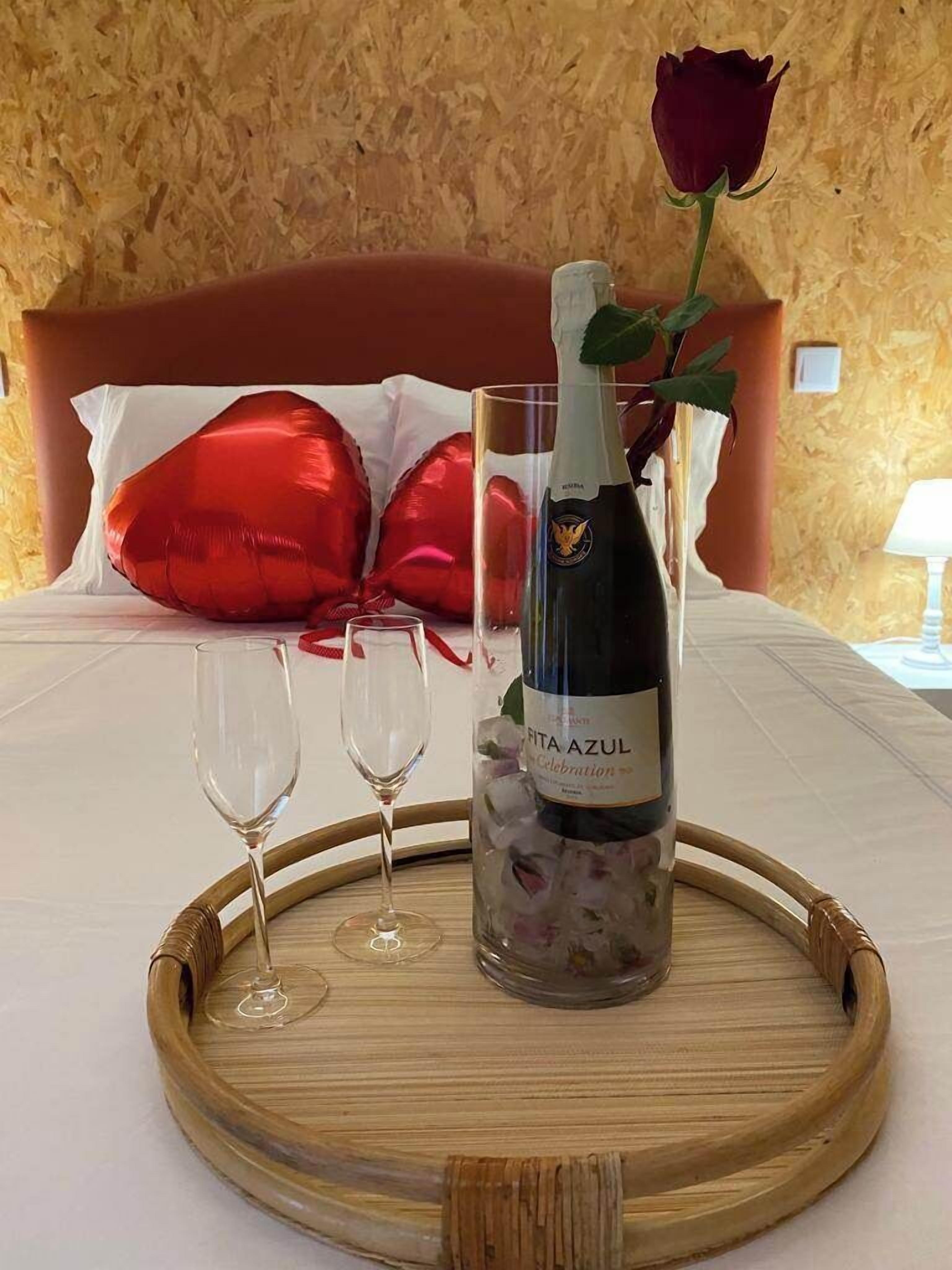 A bed with a wooden headboard, a bedside table with a bottle of Fita Azul champagne, two glasses, and a rose.