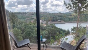 En balkong med två stolar och ett litet bord, med utsikt över en sjö och skogbeklädda kullar.