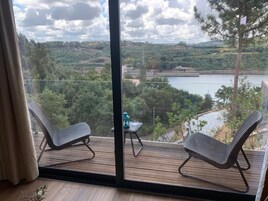 Un balcón con dos sillas y una mesita pequeña, con vistas a un lago y colinas boscosas.