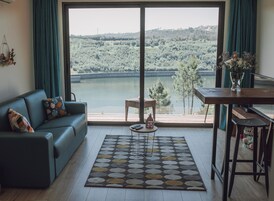 Una sala de estar con un ventanal que da a un lago y un bosque, un sofá, una mesa de centro y una mesa de comedor.
