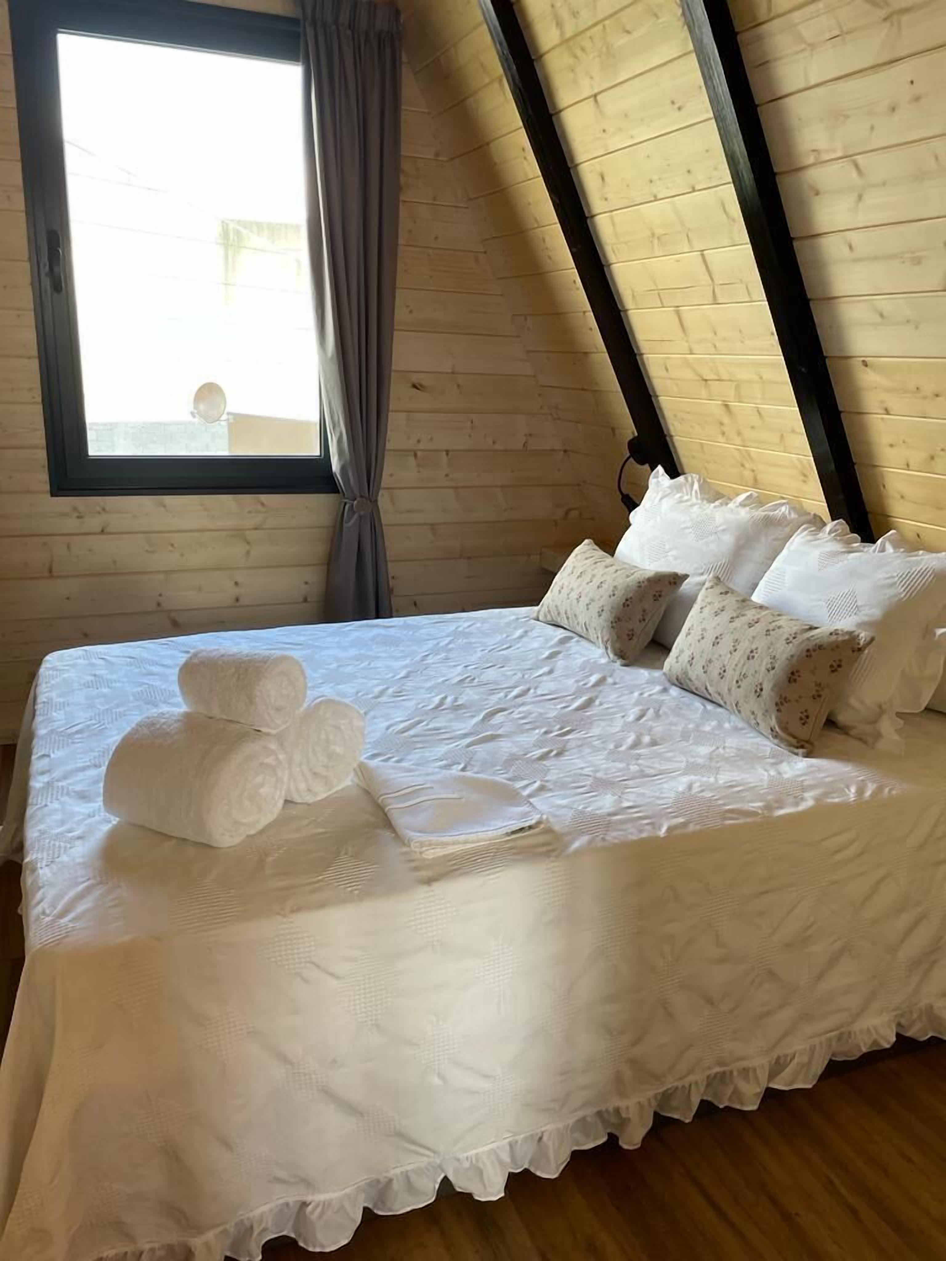 A neatly made bed with white linens and pillows, a window with curtains, and a wooden ceiling.