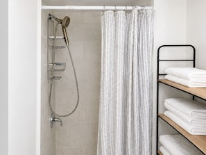A shower with a handheld showerhead, a glass shelf, and a towel rack with folded towels. - Chic Towson Townhome: 3BR, Bonus Suite, Deck, Free Parking - 15 Min to Baltimore (Towson)