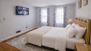 A bedroom with a bed, a TV mounted on the wall, a nightstand, and a window with curtains. - Chic Towson Townhome: 3BR, Bonus Suite, Deck, Free Parking - 15 Min to Baltimore (Towson)