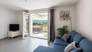 A modern living room with a blue sofa, a TV on a white cabinet, and a balcony with a table and chairs. - Magnificent T3 with all comforts, very large terrace 10 minutes walk from the beach (L'Île-Rousse)