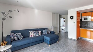 A modern living room with a blue sofa, a floor lamp, and a kitchen area with orange cabinets. - Magnificent T3 with all comforts, very large terrace 10 minutes walk from the beach (L'Île-Rousse)