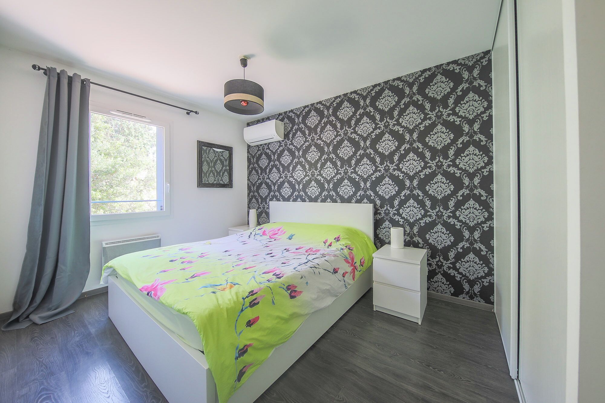 A bedroom with a bed, a bedside table, a window with curtains, a wall with a patterned wallpaper, and a ceiling light.