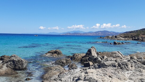 A rocky shoreline with clear blue water and distant mountains. - Magnificent T3 with all comforts, very large terrace 10 minutes walk from the beach (L'Île-Rousse)