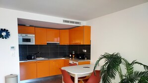 A modern kitchen with orange cabinets, a microwave, and a sink. There is a dining table with two chairs and a potted plant. - Magnificent T3 with all comforts, very large terrace 10 minutes walk from the beach (L'Île-Rousse)