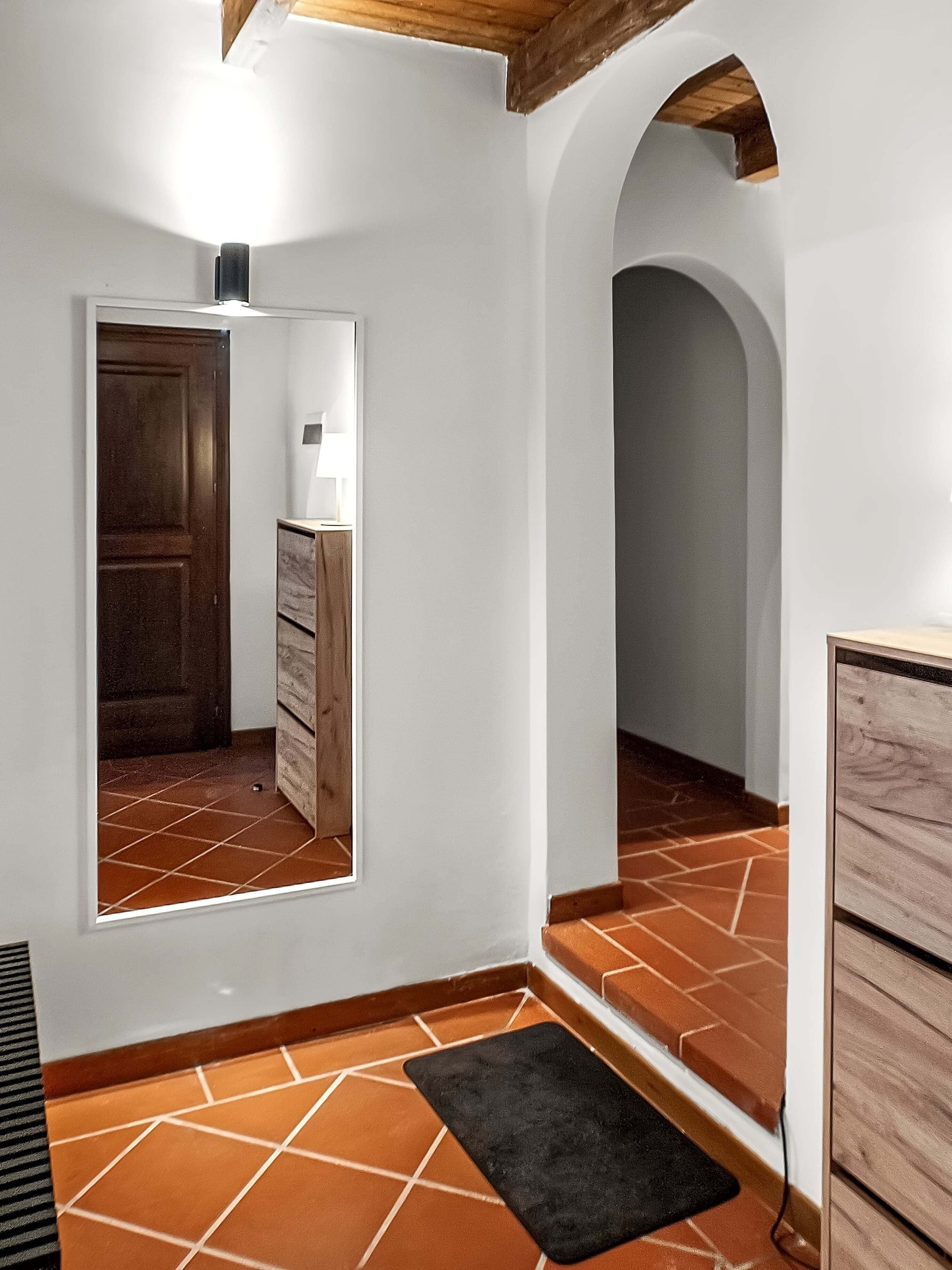 A hallway with a wooden door, a mirror, a small table with a lamp, and a black rug on a tiled floor.