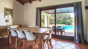 Un coin repas avec une table et des chaises en bois, une vue sur une piscine à travers de grandes portes vitrées et un sol carrelé.