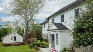 A two-story house with a stone foundation, a small porch, and a garden with various plants and flowers. - Historic 4BR/3.5BA Home near Villanova — Private Garden & Peaceful Stay
 (Villanova)