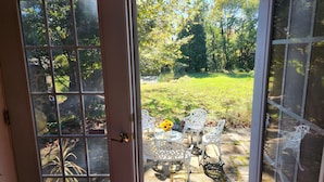 A glass door leading to an outdoor patio with white metal chairs and a table, surrounded by a grassy area and trees. - Historic 4BR/3.5BA Home near Villanova — Private Garden & Peaceful Stay
 (Villanova)