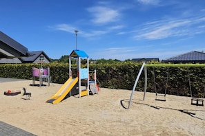 Un parque infantil con tobogán y columpios, rodeado de arena y follaje.