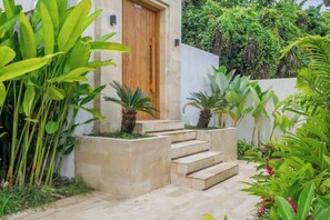 A modern building with a wooden door, a set of stone steps leading to a door, and lush greenery surrounding the entrance. - Villa Gantari by GORO (Gianyar Regency)