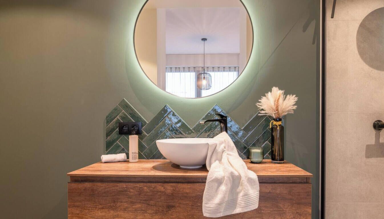 Un baño con tocador de madera, un espejo ovalado grande y salpicadero de azulejos verdes.
