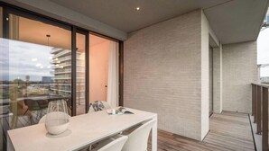 Un balcón moderno con piso de madera, una mesa blanca con un tazón y una revista, y una puerta de vidrio que da a otro balcón con vista a un edificio y un paisaje urbano.
