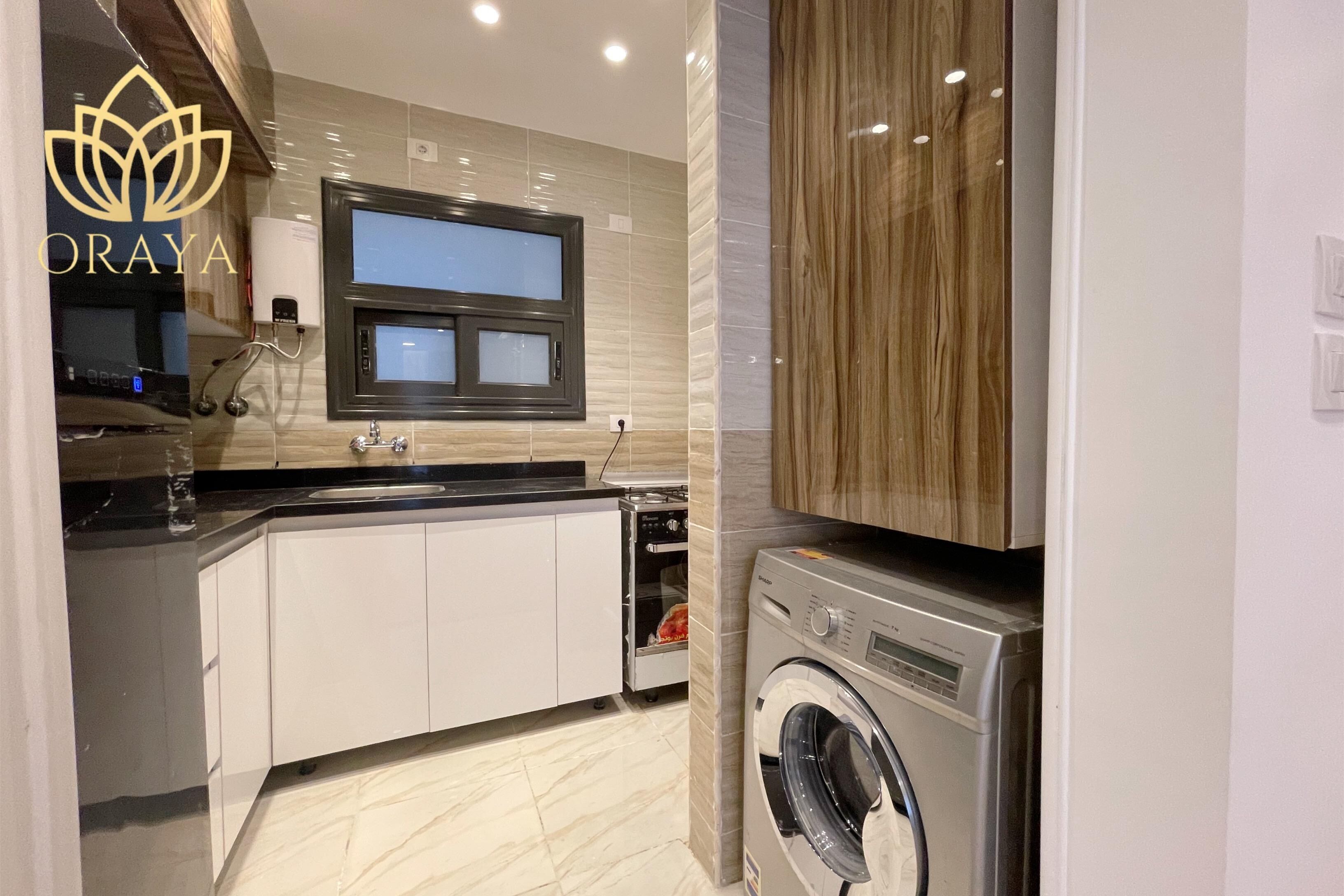 Une cuisine moderne équipée d’un lave-linge et d’un sèche-linge intégrés, d’une fenêtre au-dessus de l’évier et d’un plan de travail sur lequel se trouve un verre d’eau.