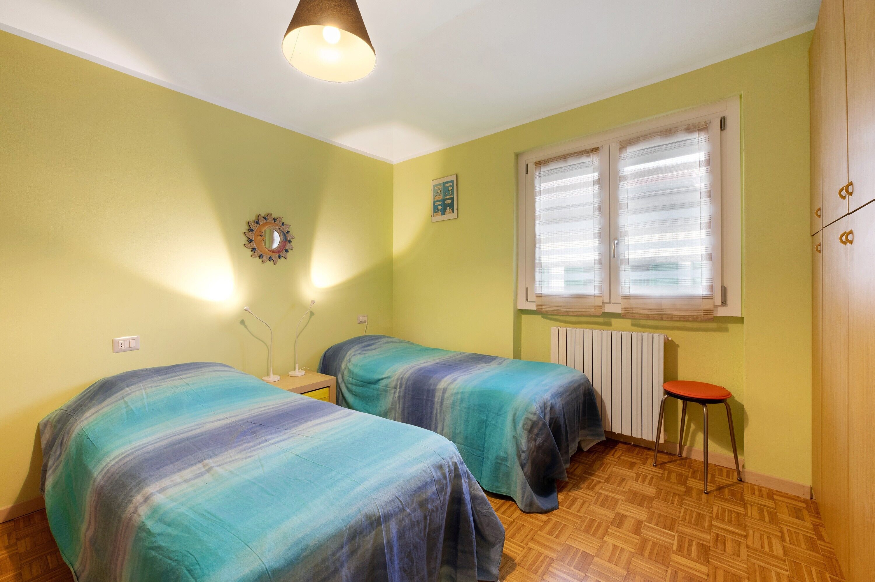 A room with two beds, a window with blinds, a radiator, a wall clock, and a small stool.