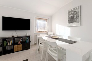 A modern living room with a flat-screen TV, a dining table with chairs, a black entertainment unit with books, and a framed photograph on the wall.