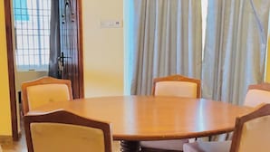 A dining area with a round wooden table surrounded by six chairs with cushioned seats. - The Tides Inn (Chennai)