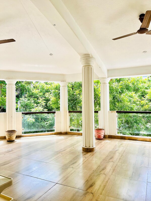 A spacious room with a high ceiling, white columns, and a view of lush greenery through large windows. - The Tides Inn (Chennai)