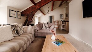 A cozy living room with a high ceiling and exposed wooden beams, a sofa set, a coffee table with a book, and a TV mounted on the wall.