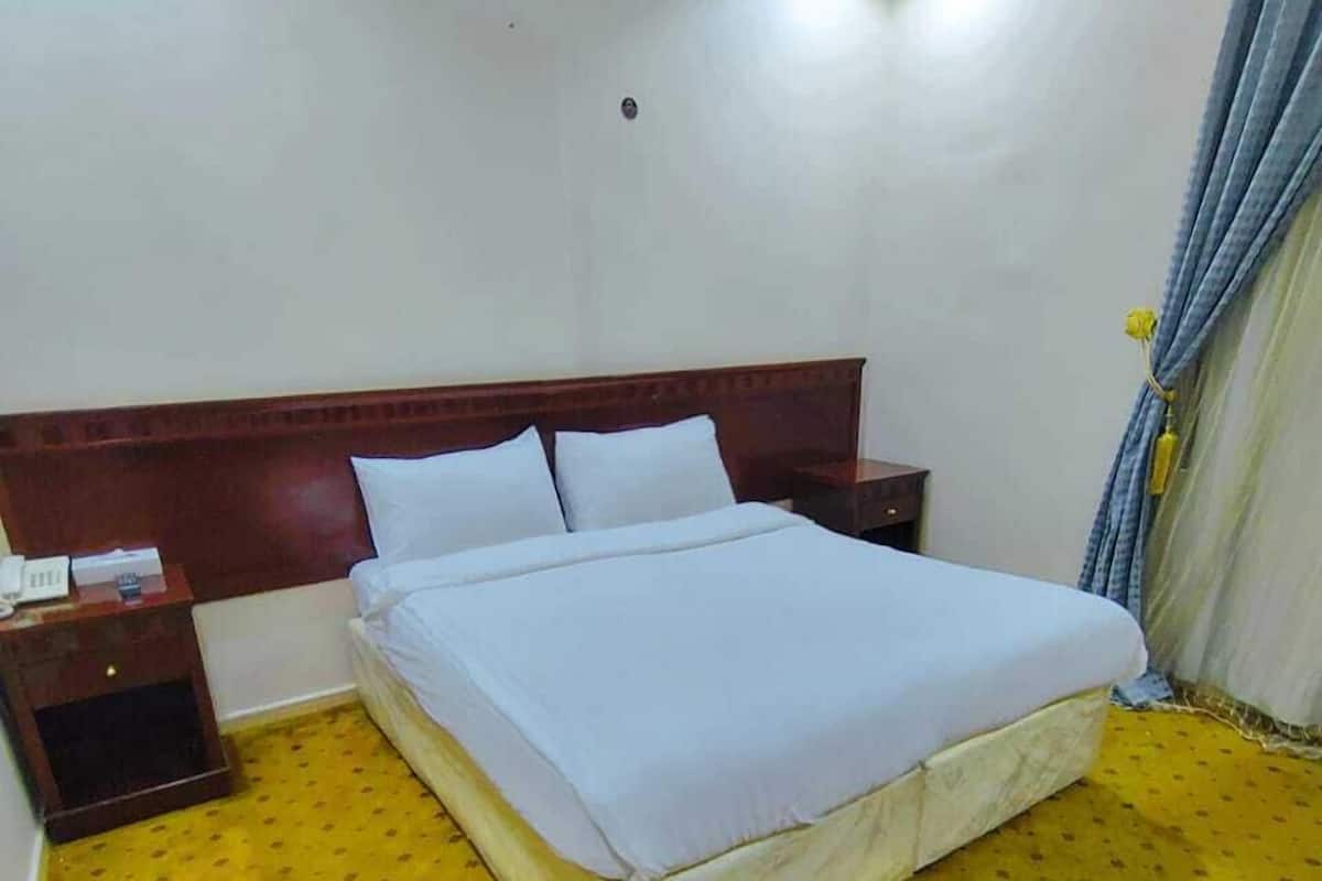 Une chambre d’hôtel avec un grand lit, une tête de lit en bois, une table de chevet et une moquette jaune à motifs.