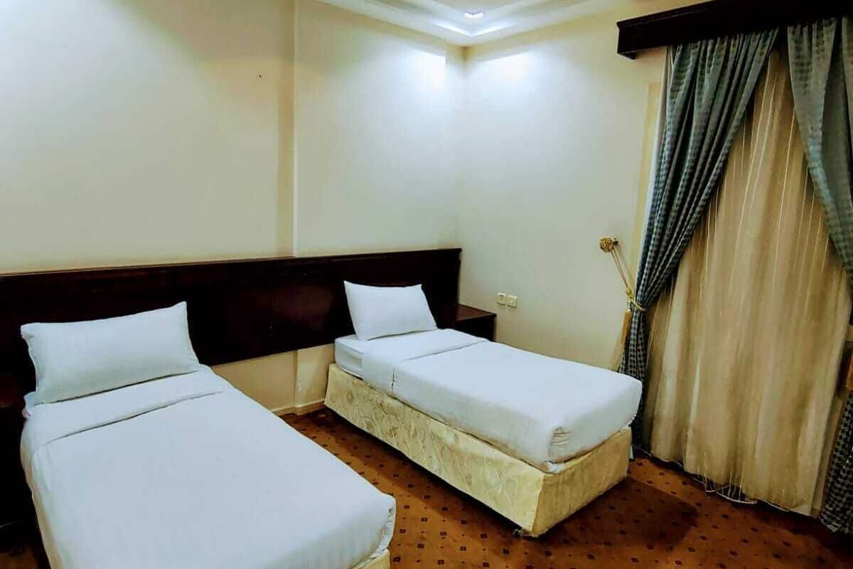 Une chambre d’hôtel avec deux lits simples, une tête de lit en bois, une grande fenêtre avec des rideaux et un sol recouvert de moquette.