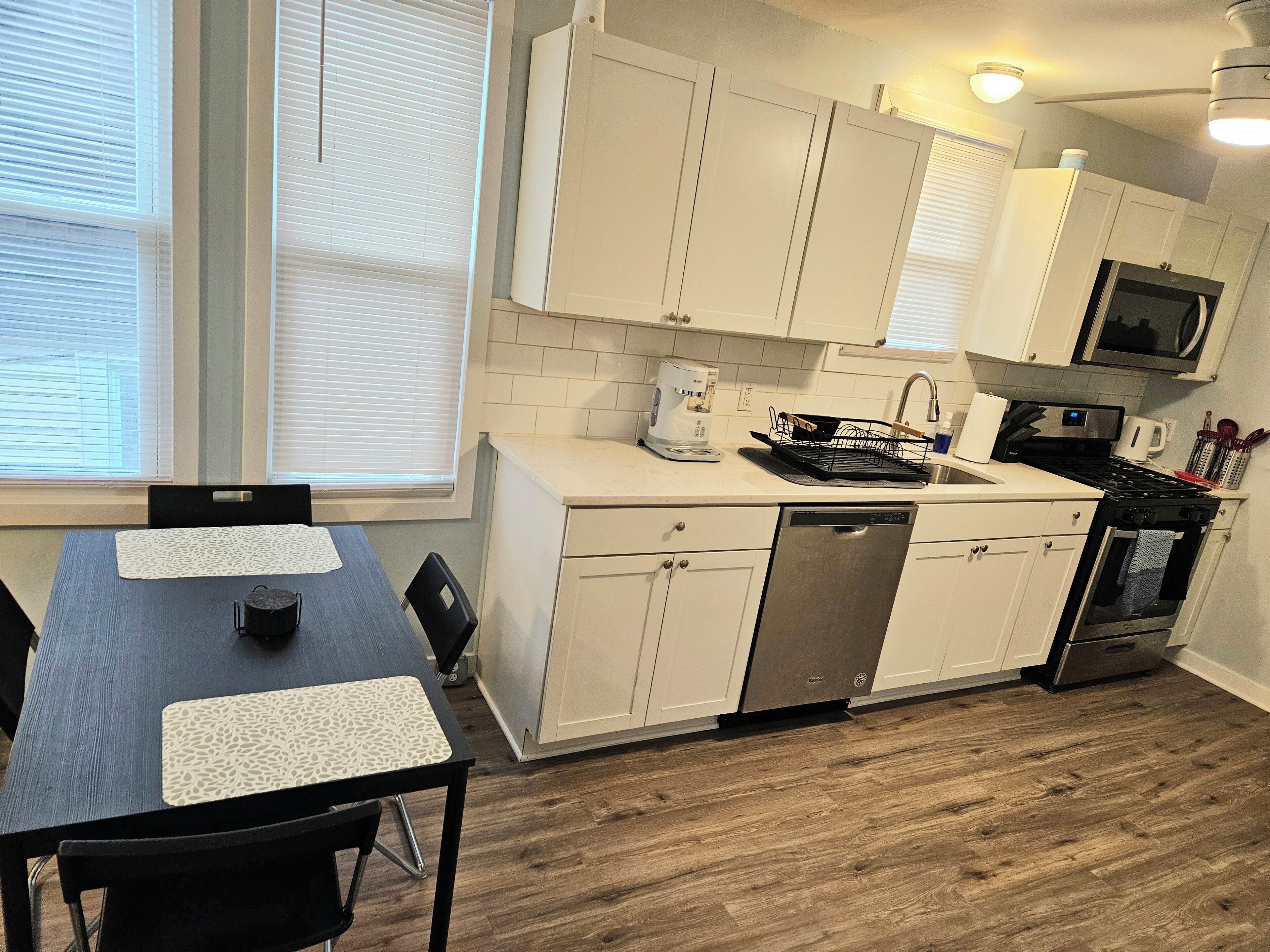 Una cocina moderna con armarios blancos, lavavajillas, estufa y microondas. Hay una mesa de comedor con sillas y una ventana con persianas.