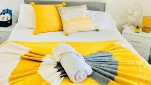 A neatly made bed with yellow and white bedding, a painting of trees on the wall, and a bedside table with a lamp and a fan. - Fratton House: Alver Road: Free parking (Fratton)