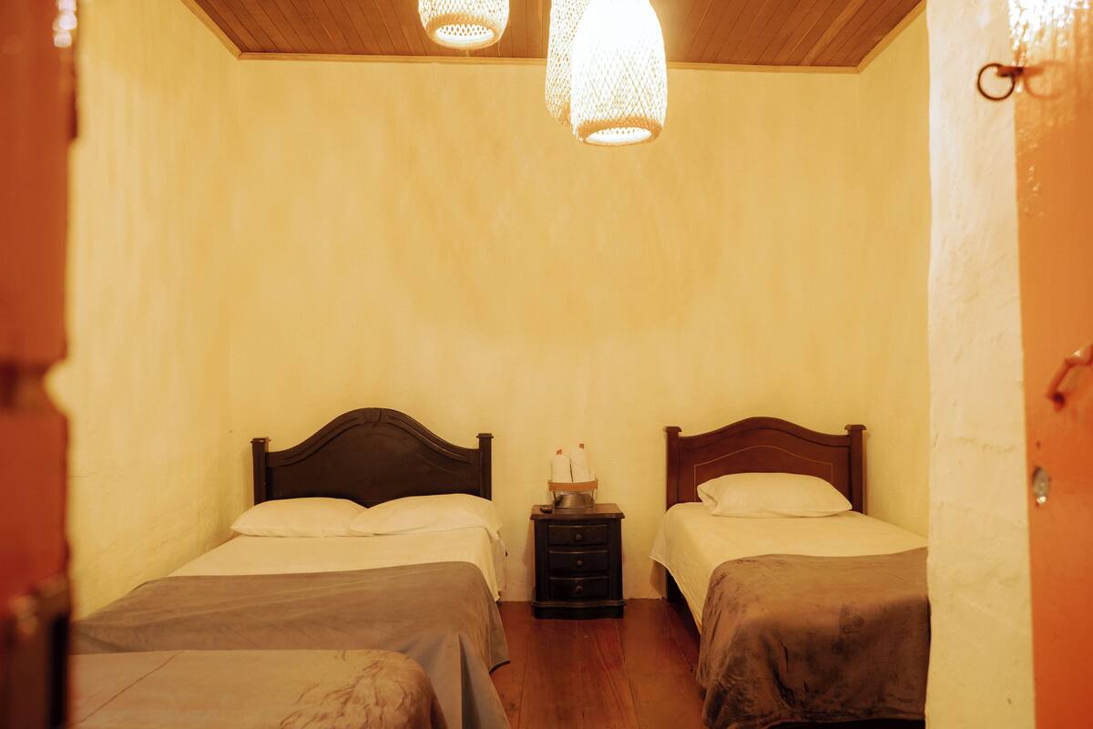 A room with two beds, wooden headboards, a nightstand with a lamp, and a wooden ceiling.