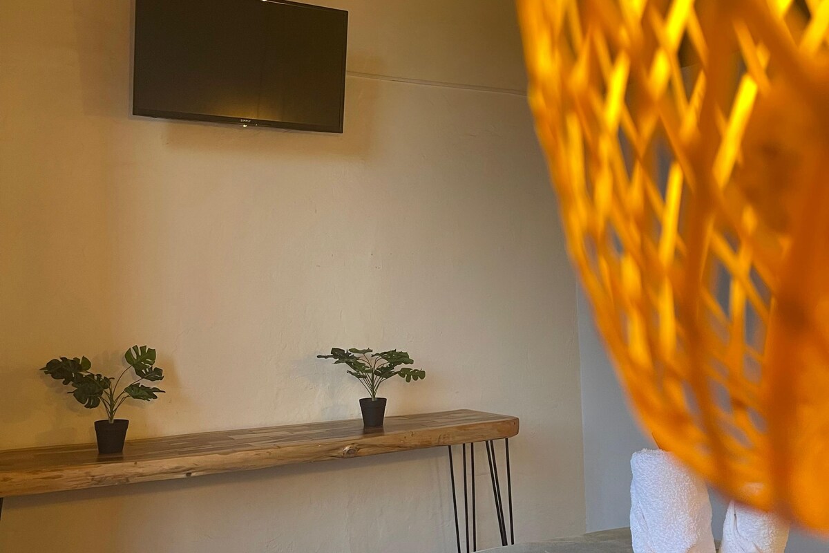 A hotel room with a bed, a TV mounted on the wall, a wooden shelf with two potted plants, and a woven basket with towels.