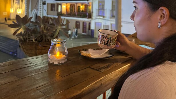 A person seated at a wooden table with a candle, a cup, and a plate. In the background, there is a building with illuminated spires and a clock tower. - Hotel Colonial Abejorral (Abejorral)