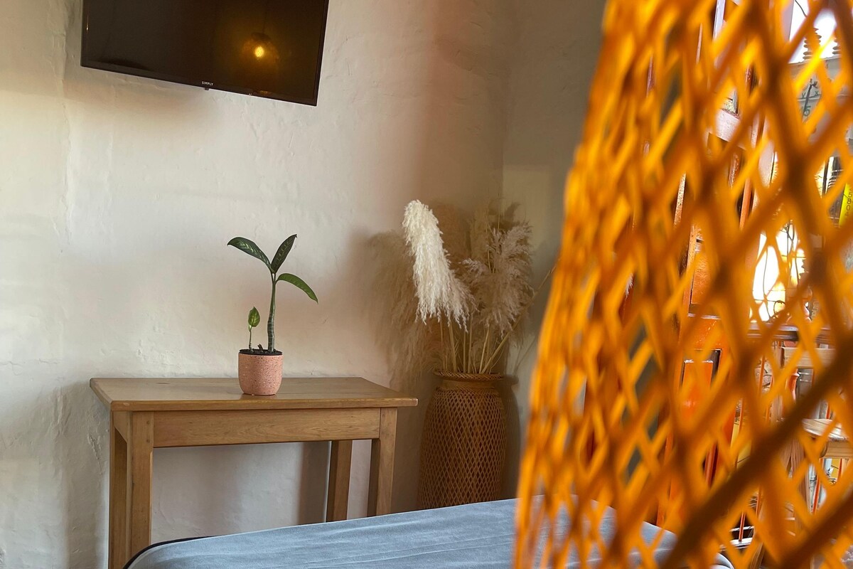 A bedroom with a bed, a small wooden table, a potted plant, and a hanging light fixture.