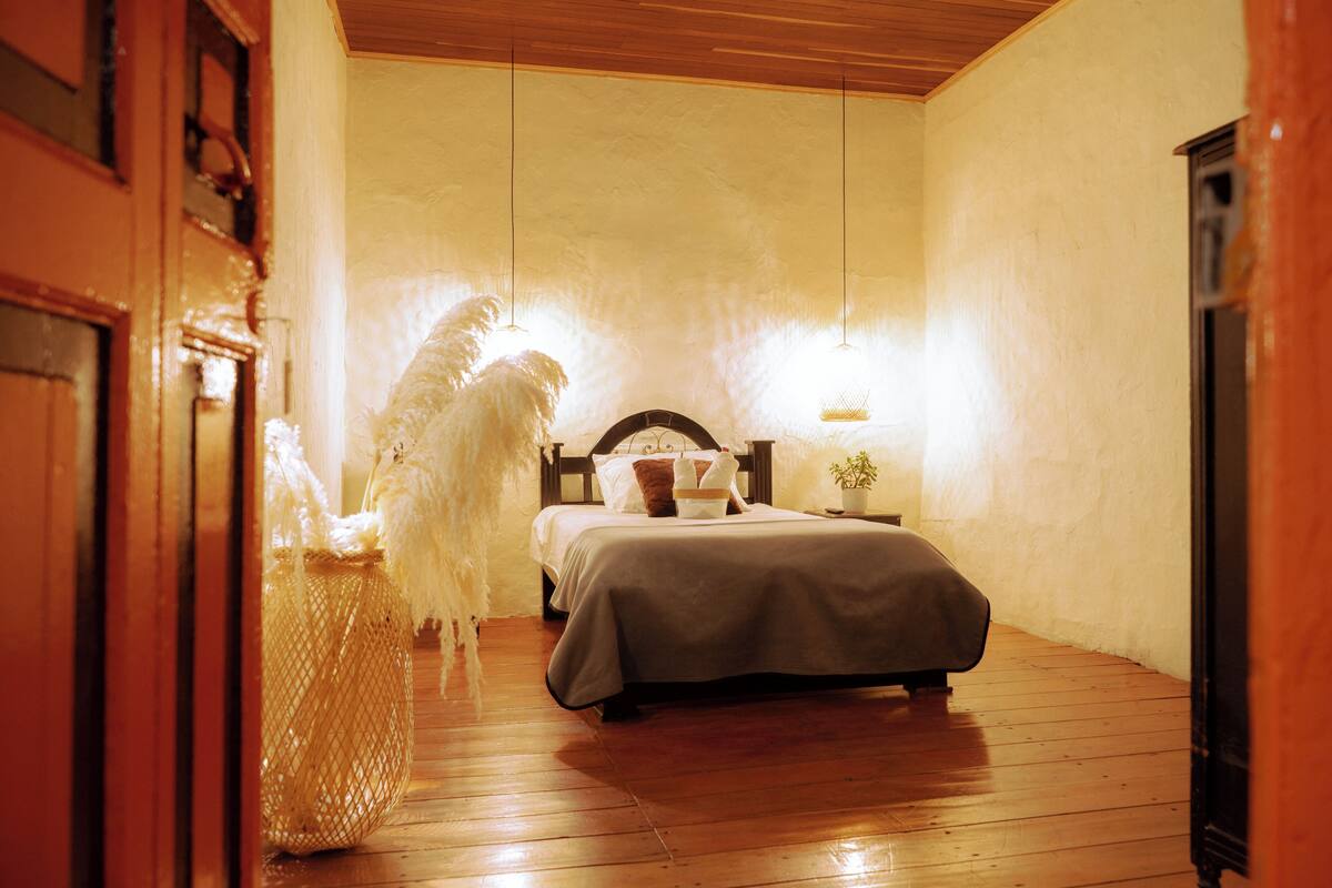 A bedroom with a wooden floor, a bed with a headboard, a bedside table, and a wall-mounted light fixture.