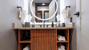 Une salle de bain avec un meuble-lavabo en bois, un miroir doté d’un éclairage circulaire et des serviettes rangées sur des étagères.