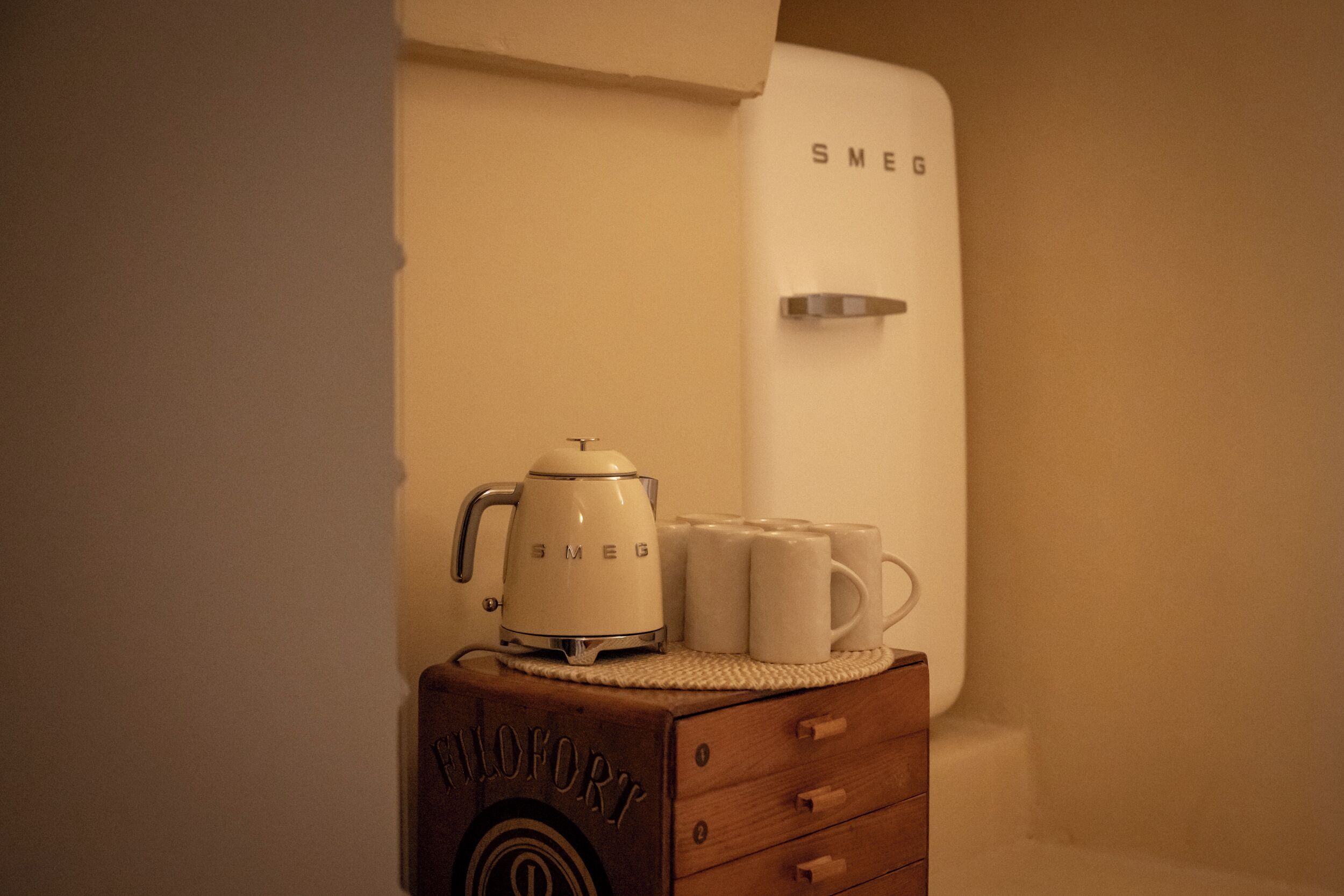 A vintage-style coffee maker and mugs on a wooden chest of drawers.