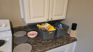 A kitchen with white cabinets, a granite countertop, and a stove. There are two gray bins on the counter, one containing food items and the other empty.