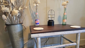 A rustic wooden table with a metal bucket on the left, containing dried flowers and a Dr. Pepper bottle.