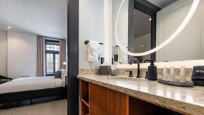 Moderni hotellihuone, jossa on sänky, puinen meikkipöytä, suuri peili sekä kylpyhuone, jossa on pesuallas ja pyyhekoukku.